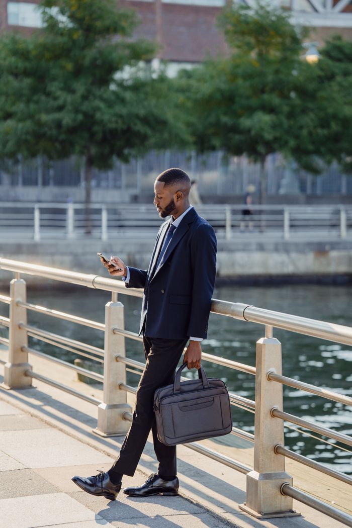 Confident businessman in a suit walking along a waterfront path checking his smartphone.