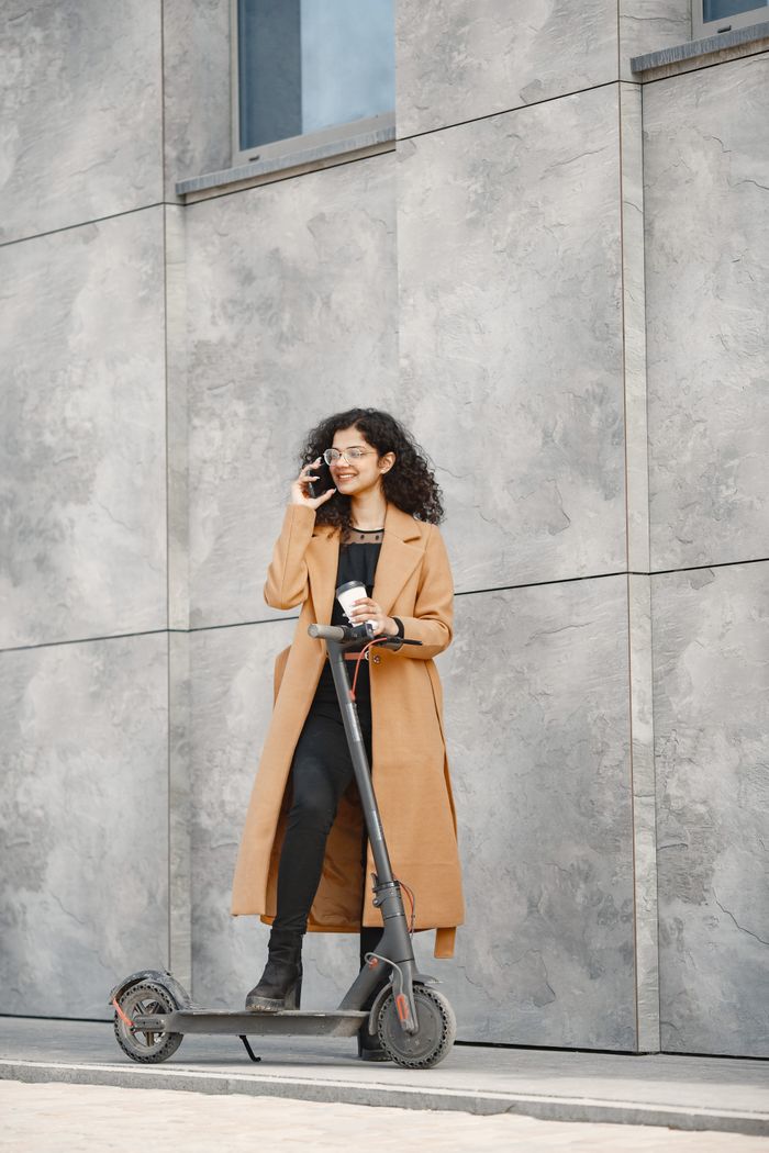 Young woman in trench coat stands with electric scooter while talking on phone outdoors.