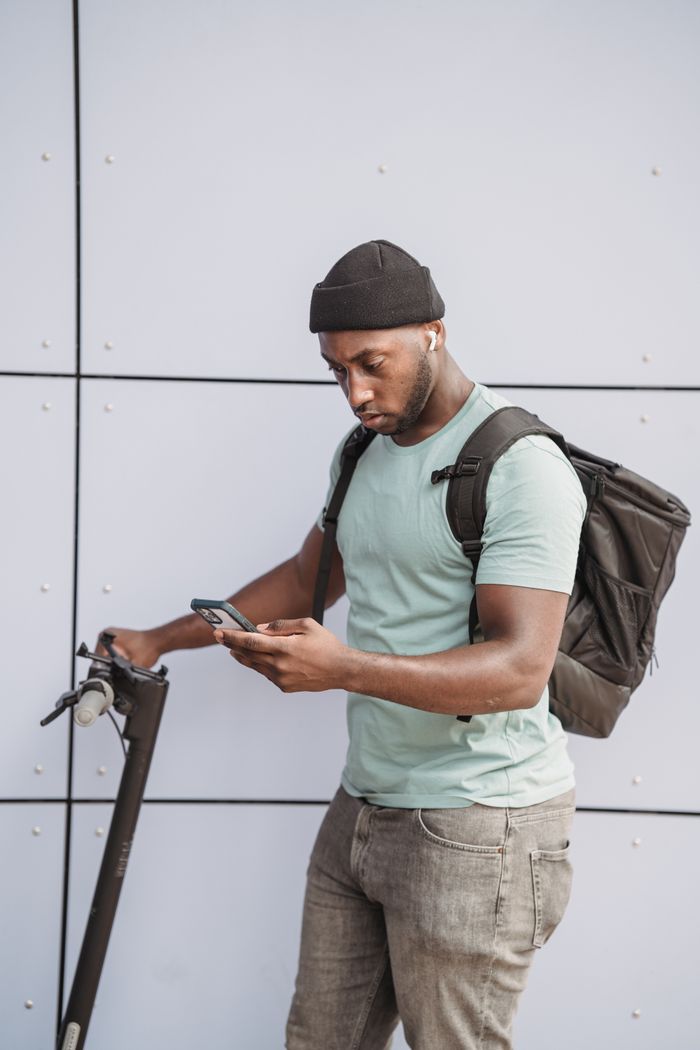 An adult man with a backpack checks his phone beside an electric scooter outdoors.